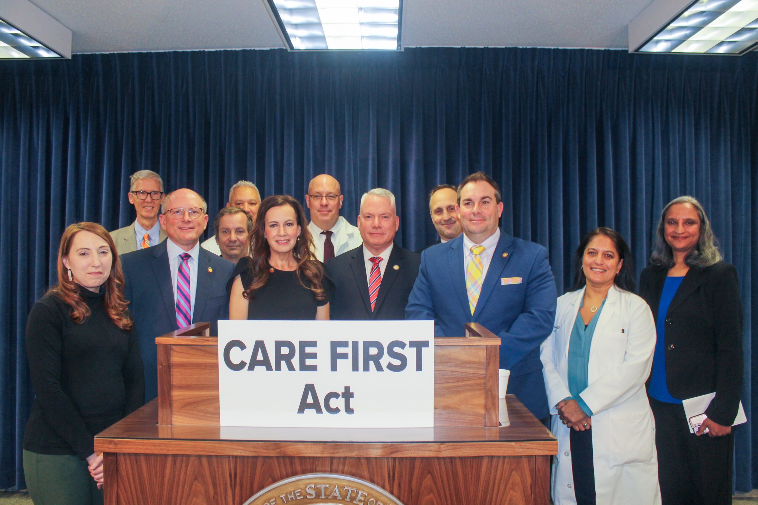 A group of physicians and lawmakers stand at a podium; sign on podium read "CARE FIRST Act"